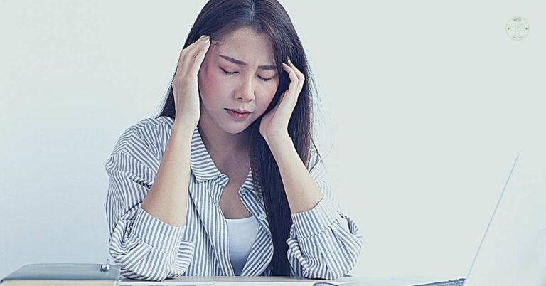 a woman holding her head in distress while working at a laptop, possibly depicting symptoms of stress-related pain