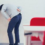 Naražená Kostrč: Příznaky & Domácí Léčba, Která Funguje! 19 a person leaning forward with hands on lower back, indicating possible pain or discomfort in the lumbar region or sacrum area.