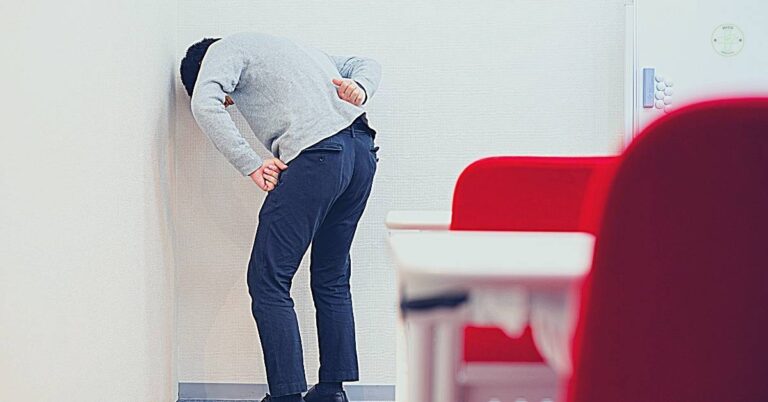 a person leaning forward with hands on lower back, indicating possible pain or discomfort in the lumbar region or sacrum area.