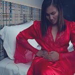 a woman in red robe sitting on bed clutching her left side near ribs, indicating abdominal discomfort or pain.