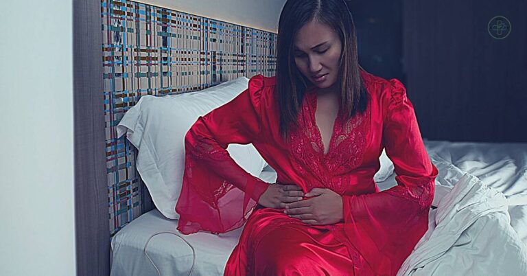 a woman in red robe sitting on bed clutching her left side near ribs, indicating abdominal discomfort or pain.