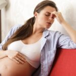 a pregnant woman holding her forehead in discomfort, sitting on a red couch in a home setting.