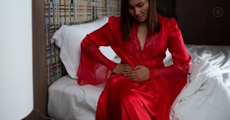 a person in red robe sitting on bed holding abdomen area, possibly indicating discomfort or pain.