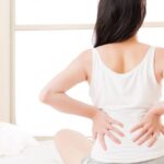 a person sitting on a bed holding their lower back and stomach area, indicating discomfort or abdominal and back pain.