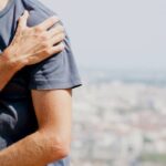 Máte Bolest Ramene Při Zvednutí Ruky? Vyzkoušejte Těchto 7 Tipů! 4 a person holding their right shoulder in pain while attempting to lift their arm, indicating shoulder discomfort or injury