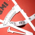 a set of measuring tapes crossed over a red background with the text "BMI" stenciled nearby, illustrating tools for calculating height and weight for healthy weight assessment