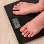 Nejpřesnější Kalkulačka Pro Výpočet Ideální Váhy Podle Výšky A Věku a Pohlaví 10 a person standing barefoot on a digital bathroom scale measuring body weight for calculating ideal weight based on height and gender.