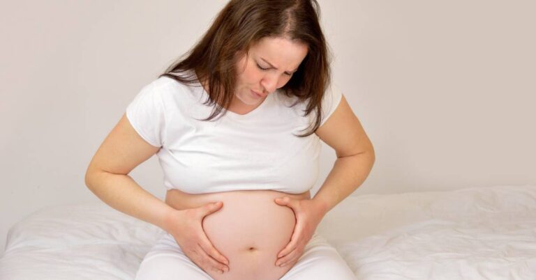 a pregnant person sitting on a bed holding her lower abdomen, showing signs of discomfort related to zvětšená děloha