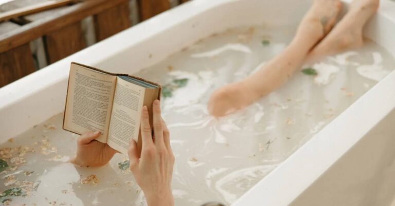 a person relaxing in a bathtub filled with bubbly water and flower petals, reading an open book while submerged up to their shoulders.