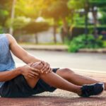 a person sitting outdoors holding their knee, showing signs of possible bolest v koleni and discomfort.