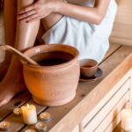 a person relaxing in a wooden sauna bench using a wooden ladle to pour water over hot stones, surrounded by candles for stress relief and muscle regeneration.