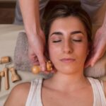 a person receiving a gentle facial massage using wooden gua sha tools, lying relaxed with hands positioned near ears