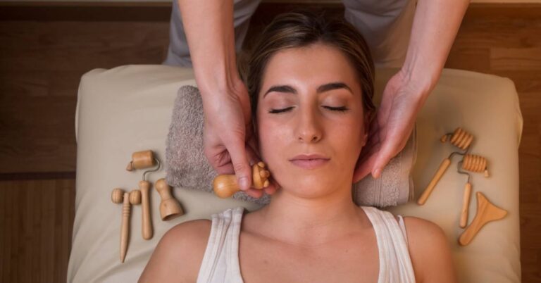 a person receiving a gentle facial massage using wooden gua sha tools, lying relaxed with hands positioned near ears