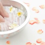 5 Nejúčinnějších Detoxikačních koupelí nohou a jak je připravit! 4 a hand gently placing white frangipani flowers into a foot soak bowl with rose petals for detoxifying foot bath relaxation