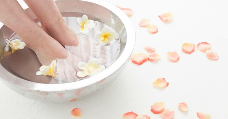 a hand gently placing white frangipani flowers into a foot soak bowl with rose petals for detoxifying foot bath relaxation