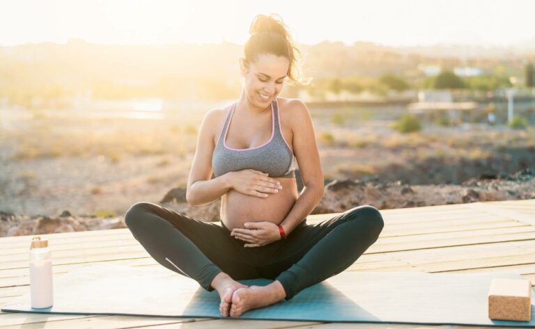 a pregnant woman practicing relaxed yoga or meditation outdoors on a wooden deck, holding her belly with a serene smile.