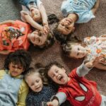 3 Relaxační techniky pro děti: Relaxace nejen pro děti s ADHD a úzkostí 8 a group of six happy children lying on their backs on a carpet, forming a circle with hands and feet, smiling and appearing relaxed together.