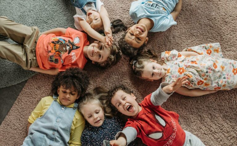 a group of six happy children lying on their backs on a carpet, forming a circle with hands and feet, smiling and appearing relaxed together.