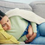 a person lying on a couch wrapped in blankets, holding their stomach while appearing uncomfortable, possibly experiencing abdominal discomfort or bloating.