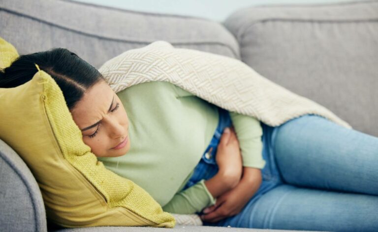 a person lying on a couch wrapped in blankets, holding their stomach while appearing uncomfortable, possibly experiencing abdominal discomfort or bloating.
